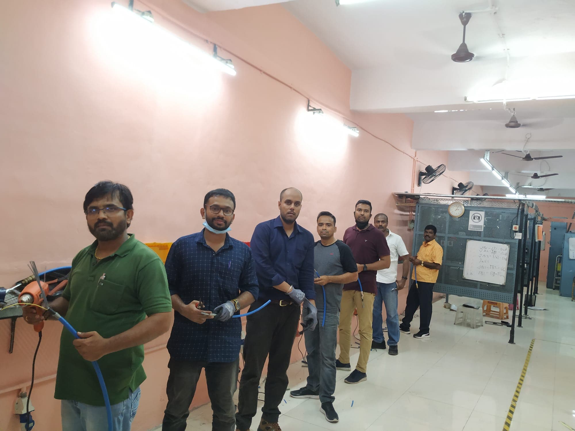 Seven engineers standing at workbenches in the training workshop, each holding a length of cable and hand tools, with a tool board and a whiteboard of cable schedules behind them.